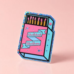 May include: A rectangular sticker featuring a matchbox design with a pink front and blue sides. The matchbox contains six matches with pink tips. A blue ribbon banner across the center displays the text Shabbat Shalom. The sticker has a holographic finish.