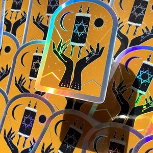 Może przedstawiać: Zestaw iryzujących holograficznych naklejek z czarno-białą ilustracją dwóch dłoni trzymających zwój ze Gwiazdą Dawida. Zwój znajduje się na żółtym tle z półksiężycem i słońcem.