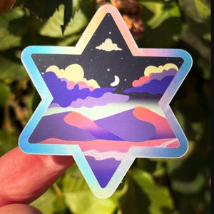 Può includere: Un adesivo a forma di stella con bordo olografico. L'adesivo presenta una scena notturna con una luna crescente, nuvole e un paesaggio di dune viola e rosa. Lo sfondo è blu scuro con stelle.