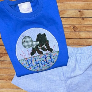 May include: A blue t-shirt and light blue shorts set. The shirt features an embroidered grasshopper design with the name "Rhett" in colorful letters. The shorts have a dotted pattern.