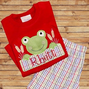 Könnte beinhalten: Rotes Kinder-Schlafanzug-Set mit einem grünen Frosch-Applikation und dem Namen "Rhett" auf einem Banner gestickt. Das Set enthält ein langärmliges Oberteil und karierte Shorts. Der Frosch hat ein Tupfenmuster und ist von Schilf umgeben.