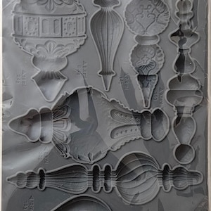 May include: A grey silicone mold by Iron Orchid Designs labeled Baubles, featuring various intricate ornament shapes. Each cavity includes small text indicating volume in ml and weight in grams, such as 18ml 24g, 8.6ml 8.9g, 8.4ml 14g, 9.3ml 14g, 21ml 32g, 12ml 18g, and 24ml 36g.