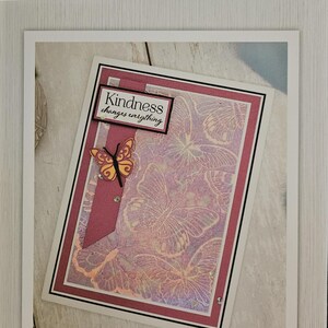 Könnte beinhalten: Ein Schmetterlings-Prägefolder von Crafter's Companion. Das Design zeigt eine Collage von Schmetterlingen mit dem Text "Kindness changes everything". Der Ordner misst 15,2 cm x 10,2 cm.