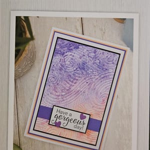 Könnte beinhalten: Ein 3D-Prägefolder mit einem Wirbelwellen-Design. Die Karte hat einen lila-rosa Farbverlaufshintergrund mit einem strukturierten Muster. Die Karte misst 15,2 cm x 10,2 cm und enthält den Text "Have a gorgeous day!"