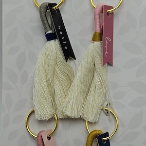 May include: Four decorative keychains with cream-colored tassels, gold rings, and tags that read "hello", "love", "thanks", and "hello". The keychains are packaged on a white and gold patterned background.