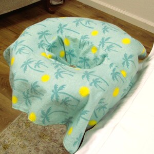 May include: A light blue massage table face cradle cover with a pattern of palm trees and yellow suns. The cover is designed to fit over the face cradle of a massage table. The cover is made of a soft, breathable fabric.