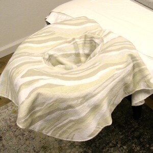 May include: A cream-colored massage table face cradle cover with a zebra stripe pattern. The cover is draped over a massage table, with a white sheet and a gray blanket visible. The room has a painting and a rug.