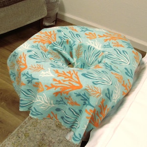 May include: A round, turquoise pouf with a coral reef pattern in orange, white, and blue. The pouf is placed on a patterned rug and wooden floor. The pouf is near a white sheet and a gray ottoman.