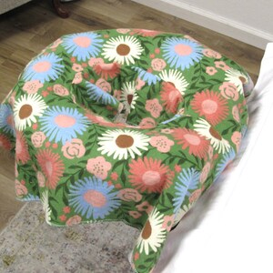May include: A round ottoman covered in a green fabric with a floral pattern. The pattern includes large flowers in shades of pink, blue, and white, with brown centers and green leaves. The ottoman is placed on a light-colored rug.