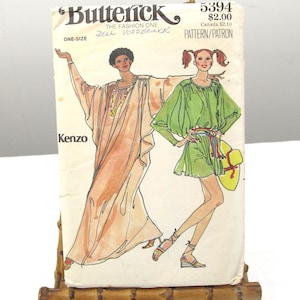 May include: A vintage Butterick 5394 sewing pattern envelope featuring illustrations of two garments: a long, flowing peach-colored kaftan and a short, bright green tunic dress. The envelope displays the Kenzo brand name and is held by a bamboo stand.