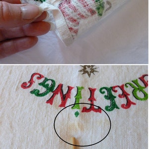 May include: White cotton tea towel with a red, green, and gold Christmas design. The towel has the words "Greetings" and "Holiday" printed on it. There is a small yellow stain on the towel.