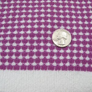 K&ouml;nnte beinhalten: Lilafarbener und wei&szlig;er Strickstoff mit geometrischem Muster. Eine US-Viertel-Dollar-M&uuml;nze liegt auf dem Stoff zur Gr&ouml;&szlig;enbestimmung.