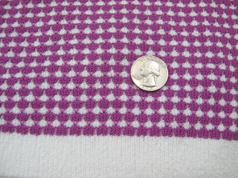 K&ouml;nnte beinhalten: Lilafarbener und wei&szlig;er Strickstoff mit geometrischem Muster. Eine US-Viertel-Dollar-M&uuml;nze liegt auf dem Stoff zur Gr&ouml;&szlig;enbestimmung.