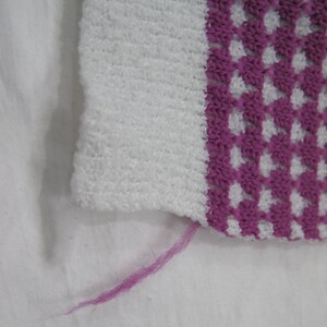 K&ouml;nnte beinhalten: Ein wei&szlig;er und rosa gestrickter Stoff mit einem rosa Faden, der vom Rand h&auml;ngt.
