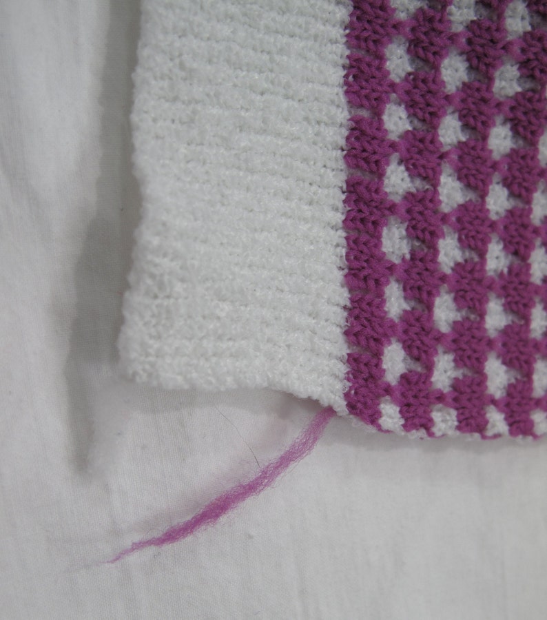 K&ouml;nnte beinhalten: Ein wei&szlig;er und rosa gestrickter Stoff mit einem rosa Faden, der vom Rand h&auml;ngt.