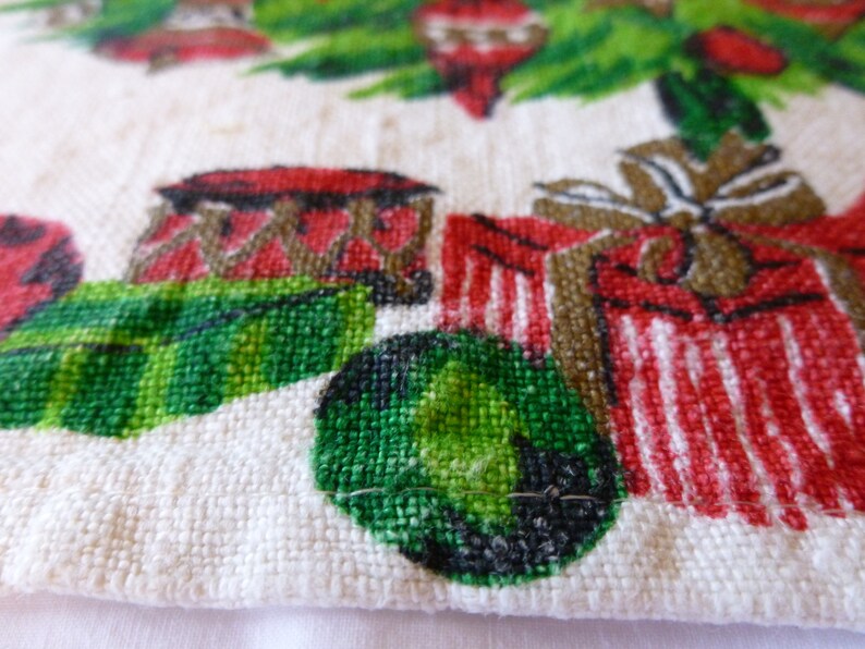 May include: A white cotton tea towel with a Christmas-themed design. The design features a green Christmas tree with red and white ornaments, red and white striped presents, and a green and black ball.