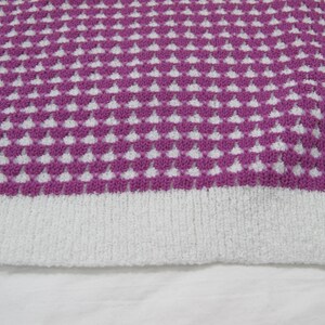 K&ouml;nnte beinhalten: Nahaufnahme eines gestrickten Stoffes mit einem wei&szlig;en und violetten geometrischen Muster. Der Stoff hat eine weiche, flauschige Textur.