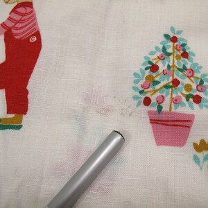 May include: White fabric with a red and green illustration of a person holding a star and a pink and green tree with red and orange fruit in a pink pot.