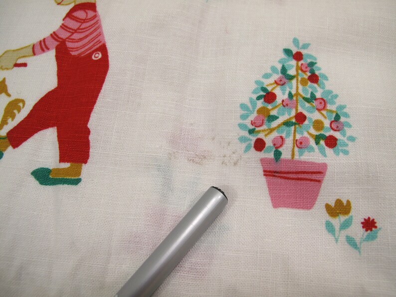 May include: White fabric with a red and green illustration of a person holding a star and a pink and green tree with red and orange fruit in a pink pot.