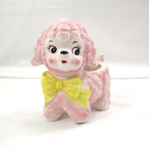 May include: A pink ceramic poodle figurine with a yellow bow. The poodle has a curly coat, black eyes, and a red mouth. The figurine is a planter, with an opening on its back.
