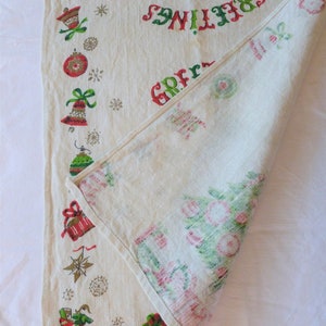 May include: A white linen tablecloth with a red and green Christmas tree design. The tablecloth has a border of Christmas ornaments, bells, and stars. The text "Greetings" is printed in red and green on the tablecloth.