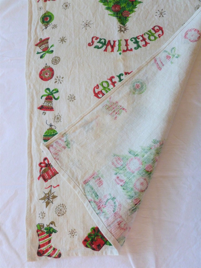 May include: A white linen tablecloth with a red and green Christmas tree design. The tablecloth has a border of Christmas ornaments, bells, and stars. The text "Greetings" is printed in red and green on the tablecloth.