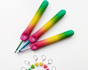 May include: Three crochet hooks with rainbow-colored handles and metal tips, along with six heart-shaped stitch markers. The handles transition from green to yellow to pink. The stitch markers are in various colors.