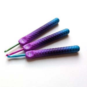 May include: Three crochet hooks with gradient purple, blue, and teal handles. The handles have a scale-like texture. The hooks are numbered 4, 4.5, and 5. The hook tips are green, pink, and blue.