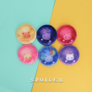 Könnte beinhalten: Sechs runde, bunte, flauschige Stoff-Patches mit einem Katzenkopf-Design. Jeder Patch hat eine andere Farbe und ein anderes Katzenkopf-Design. Die Patches sind in einem Kreis auf einem blauen und gelben Hintergrund angeordnet. Der Text "S.P.O.I.L.E.R." ist auf dem gelben Hintergrund gedruckt.
