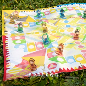 May include: A colorful game board with a yellow, blue, and green design. The board features a variety of characters, including a girl with brown hair, a boy with blonde hair, and a girl with pink hair. The board also features the text "S.P.O.I.L.E.R."