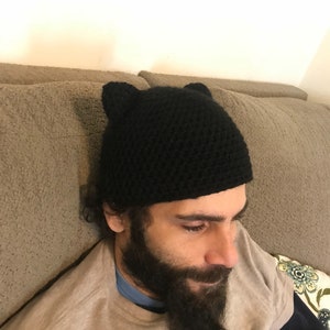 May include: A black crocheted beanie with cat ears. The beanie is worn by a person with a beard.