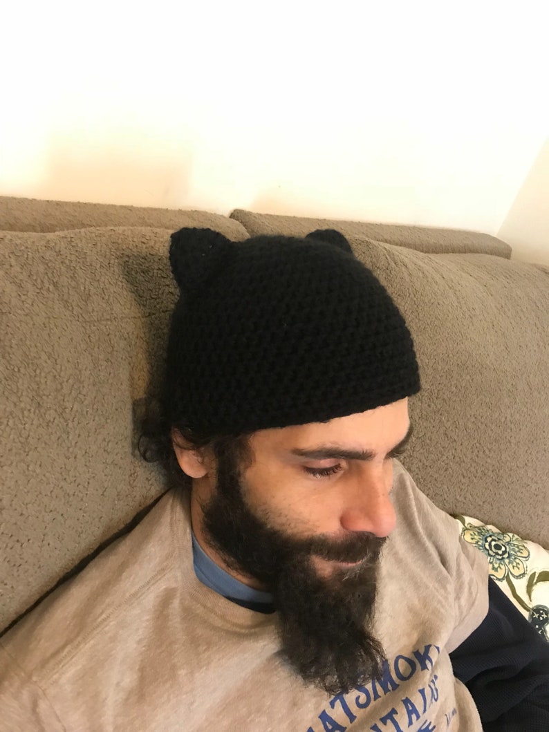 May include: A black crocheted beanie with cat ears. The beanie is worn by a person with a beard.