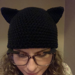 May include: A black crocheted cat ear beanie with a person wearing glasses underneath.
