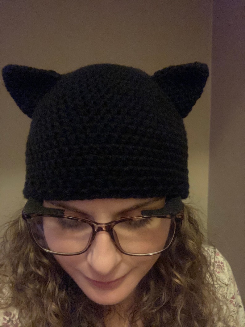 May include: A black crocheted cat ear beanie with a person wearing glasses underneath.
