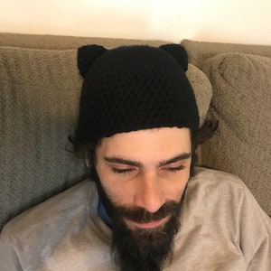 May include: A black crocheted beanie with cat ears. The beanie is worn by a person with a beard.