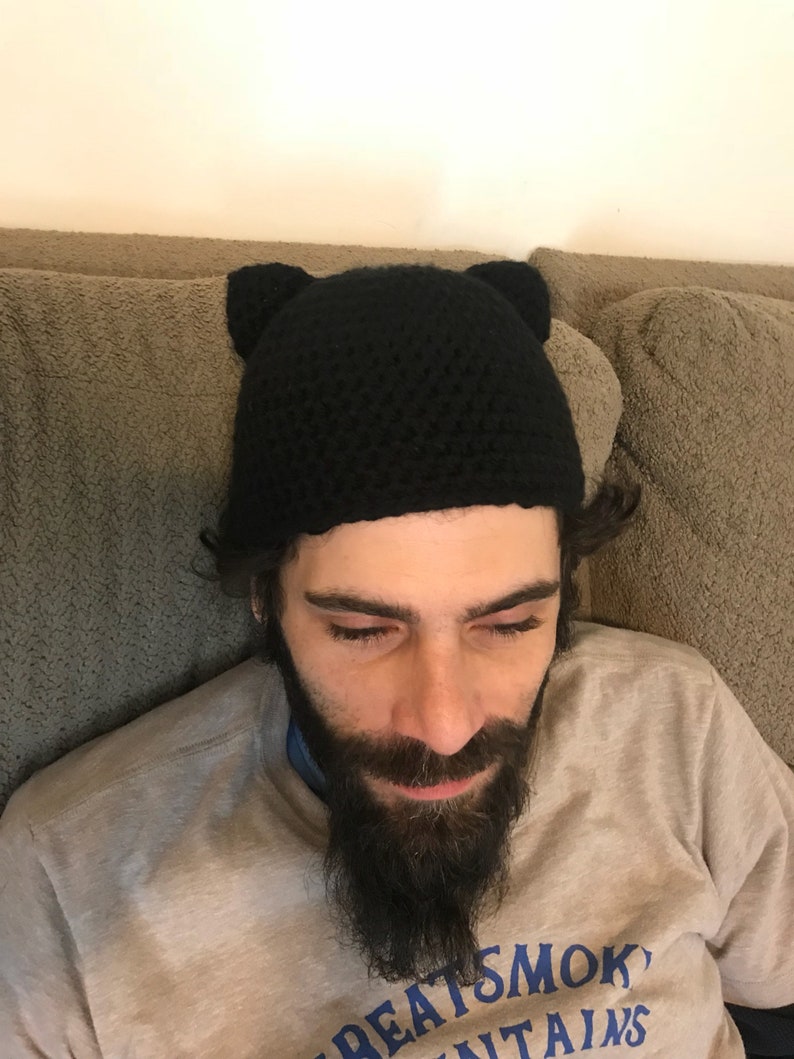 May include: A black crocheted beanie with cat ears. The beanie is worn by a person with a beard.