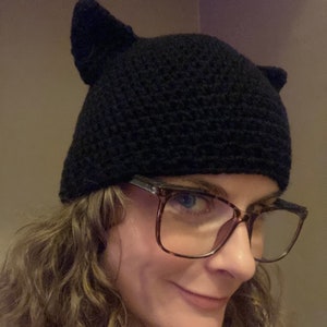 May include: A black crocheted cat ear beanie with a person wearing brown tortoiseshell glasses.