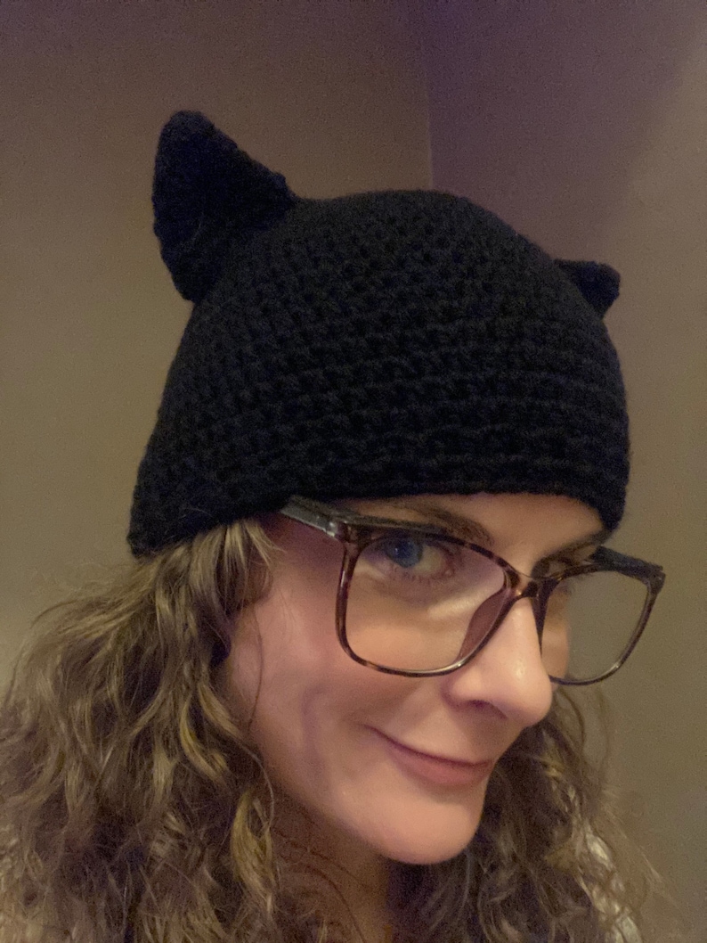 May include: A black crocheted cat ear beanie with a person wearing brown tortoiseshell glasses.