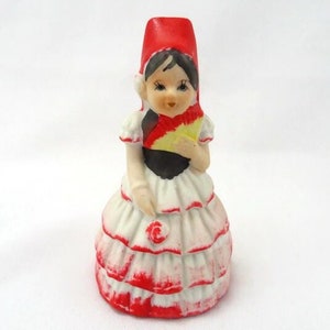 May include: A ceramic figurine of a woman in traditional Spanish attire. The figurine features a red hat, a white dress with red trim, and a fan with yellow and red accents. The figure has black hair and is holding a fan.