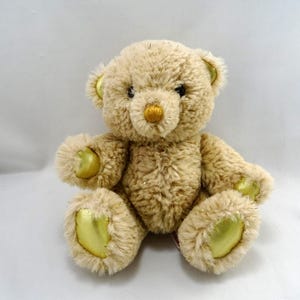 Peut inclure: Un ours en peluche avec une fourrure marron clair et des accents dorés sur les oreilles, les pattes et le nez. L'ours est assis, mettant en valeur sa texture douce et câline. Un jouet classique pour enfants.