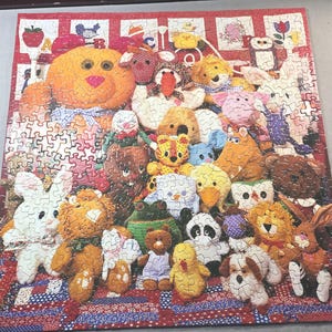 May include: A completed jigsaw puzzle featuring a colorful array of plush toy animals. The puzzle includes a large orange bear, a white rabbit, a panda, and many other animal characters. The puzzle has a red border with a patchwork design.