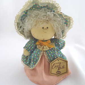 May include: A vintage doll with a fabric body, wearing a blue patterned dress and a matching bonnet with lace trim. The doll has a peach-colored skirt and a tag that reads "Orgel Doll". The doll has white curly hair and a yellow bow.