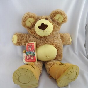 May include: A plush brown bear toy with a light yellow belly and matching boots. The bear has a brown nose and a tag that reads "Furskins". The bear is lying on a white surface.