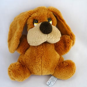 May include: A plush brown dog toy with long floppy ears, a tan muzzle, and a dark brown nose. The dog has yellow eyes and is sitting upright. A white tag is attached to the toy.