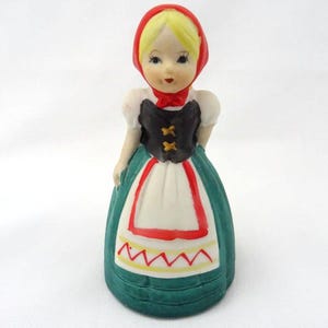 May include: A ceramic figurine of a girl in traditional attire. She wears a red head covering, a black bodice with gold accents, a white apron with red trim, and a green skirt. The figurine has blonde hair and a painted face.