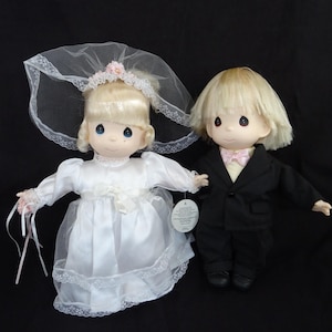 May include: Two Precious Moments dolls, a bride and groom, are displayed together. The bride doll wears a white satin dress with a lace overlay and a veil. The groom doll is in a black tuxedo with a pink bow tie. Both dolls have blonde hair.