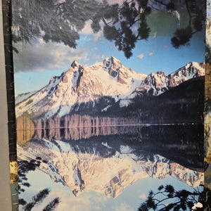 May include: A jigsaw puzzle featuring a scenic mountain landscape reflected in a calm lake. The snow-capped peaks are framed by dark green trees and a blue sky with clouds. The puzzle pieces are visible around the edges.