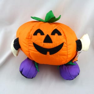 May include: A plush pumpkin toy featuring a bright orange body with a black felt jack-o'-lantern face, green fabric leaves on top, white fabric hands with black cuffs, and two round purple fabric feet attached to the bottom of the pumpkin.