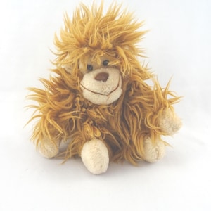 May include: A plush toy monkey with shaggy, light brown fur. The stuffed animal has a smiling face with a tan nose and a cream-colored mouth. Its arms and feet are also cream-colored. The toy is sitting on a white surface.