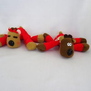 May include: Two plush reindeer toys with brown bodies, red legs, and tan hooves. The reindeer have black button noses, brown antlers, and red bows. The toys are lying on a white surface.